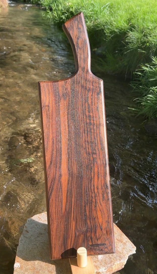A rectangular wooden cutting board with a curved handle, made from dark reddish-brown wood with prominent grain, standing on a stone block in a stream outdoors.